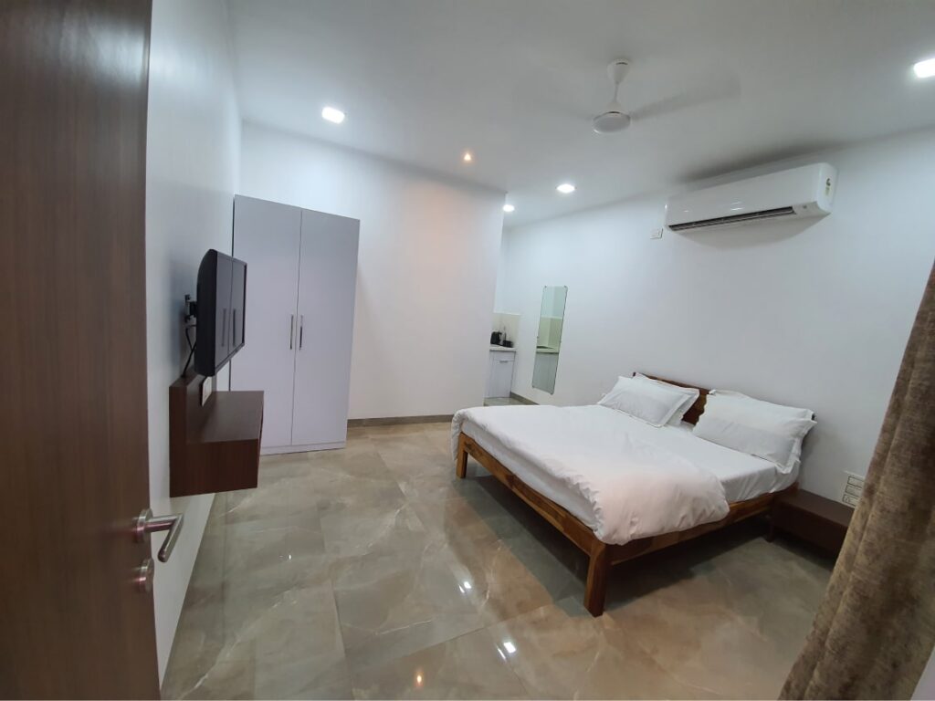 luxurious room with queen size bed ,white bedsheet ,white almirah ,AC ,fan and tv mounted on the wall.